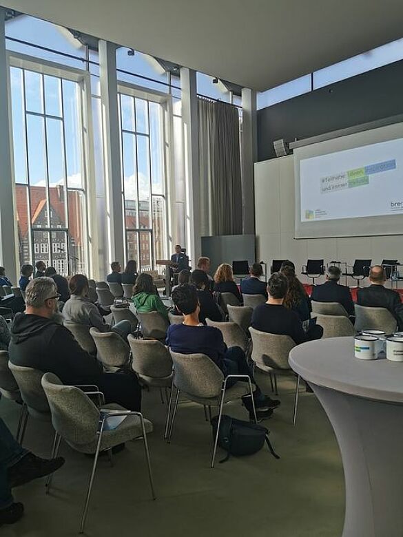 Blick aus dem voll besetzten Saal heraus auf das Podium, Arne Frankenstein spricht