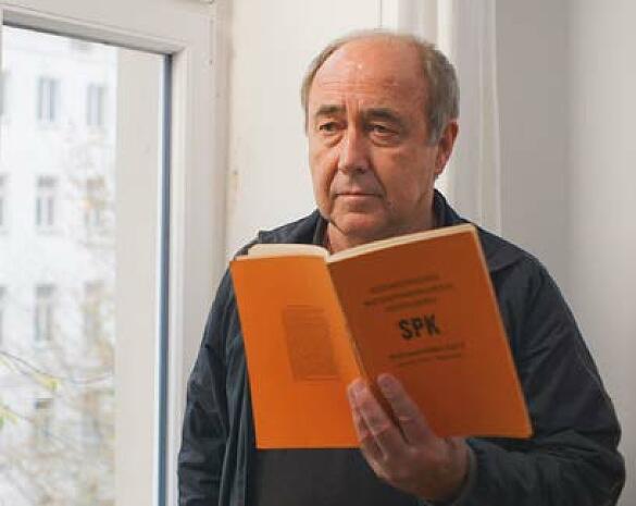 Filmszene: Ein traurig dreinblickender Mann mit Halbglatze hält ein oranges Buch mit dem Titel SPK